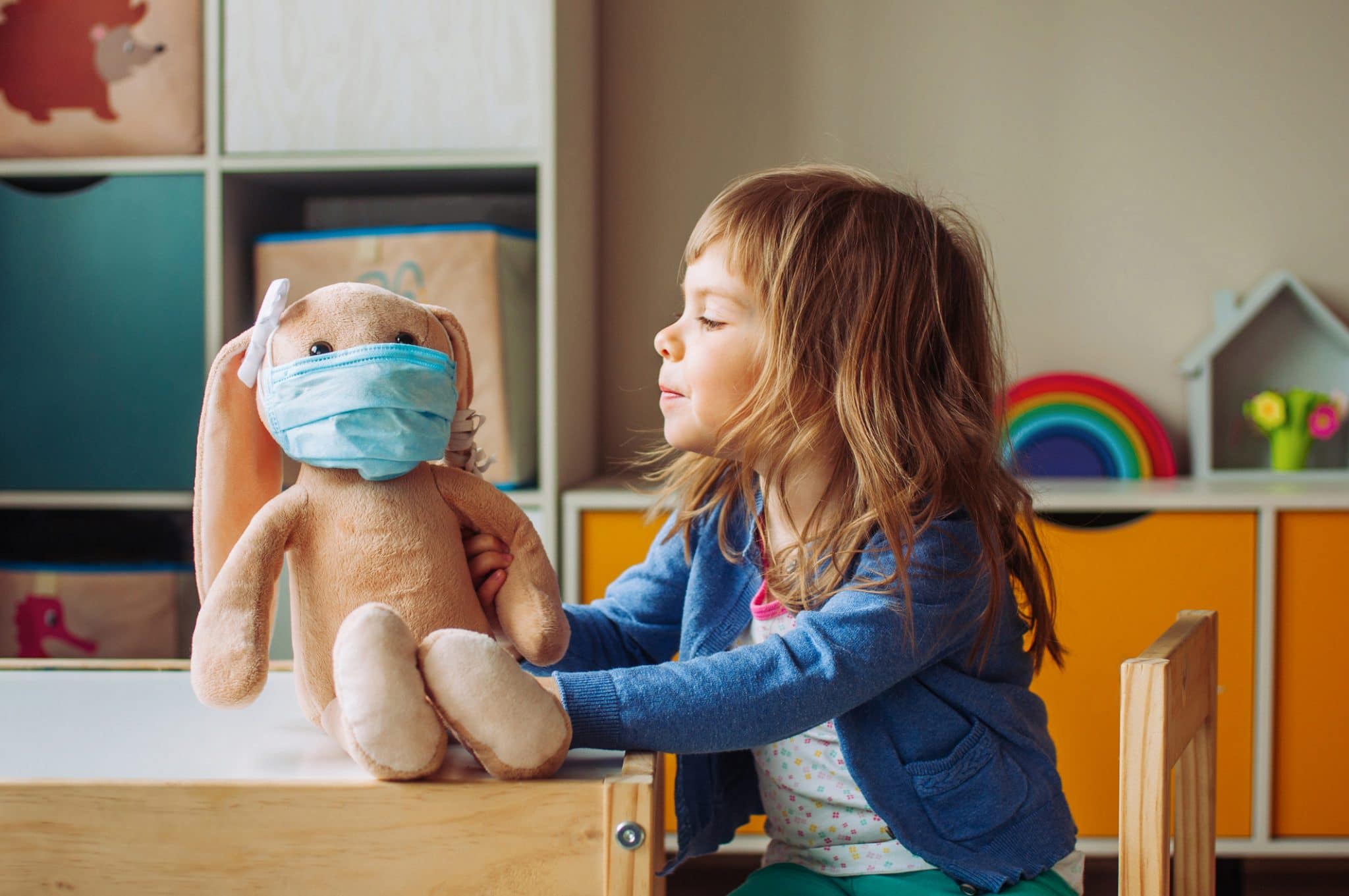 Little girl playing with rabbit soft toy in the medicine mask sitting at the table in the kids room.